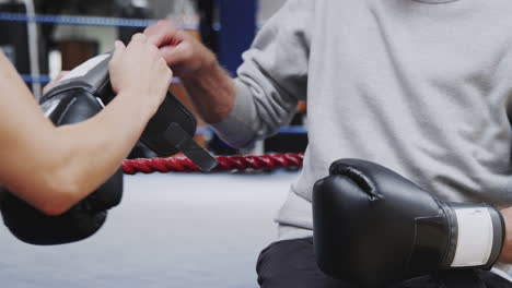 Female-Personal-Trainer-Helping-Senior-Male-Boxer-Putting-Boxing-Gloves-On-In-Gym