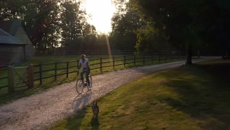 Toma-De-Drone-De-Una-Mujer-Con-Un-Perro-Mascota-Montando-En-Bicicleta-Por-Un-Camino-Rural-Al-Atardecer