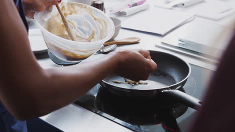 Close-Up-Of-Female-Teacher-Adding-Flatbread-Mixture-To-Pan-In-Cookery-Class