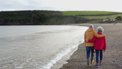 Drone-Shot-Of-Senior-Couple-Arm-In-Arm-From-Behind-Walking-Along-Shoreline-On-Winter-Beach-Vacation