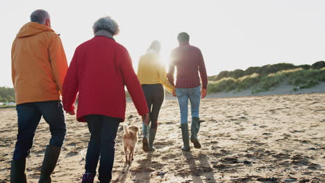 Senior-Couple-Hold-Hands-As-They-Walk-Along-Beach-With-Adult-Offspring-On-Winter-Vacation