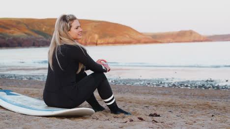 Rear-View-Of-Woman-Wearing-Wetsuit-Sitting-On-Surfboard-And-Looking-Out-To-Sea