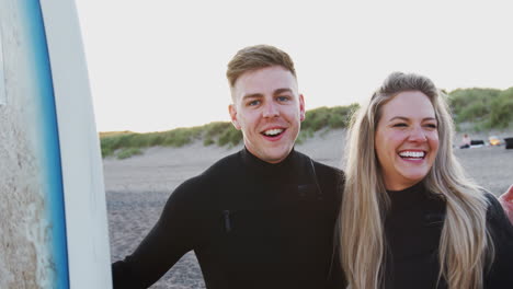 Portrait-Of-Smiling-Young-Couple-Wearing-Wetsuits-Enjoying-Surfing-Vacation-On-Beach