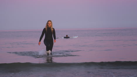 Couple-Wearing-Wetsuits-Surfing-Together-As-Woman-Walks-Out-Of-Sea