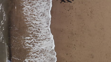 Drone-Shot-Of-Family-On-Vacation-Running-Along-Beach-By-Breaking-Waves