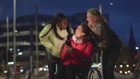 Woman-In-Wheelchair-Having-Night-Out-With-Friends-In-City-Looking-At-Mobile-Phone-Together
