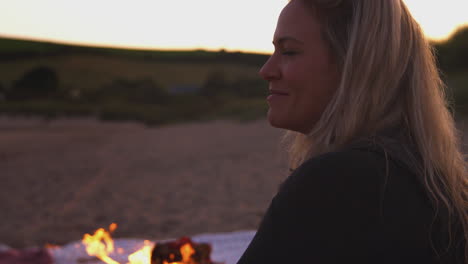 Woman-Sitting-By-Camp-Fire-On-Beach-As-Sun-Sets-Behind-Her