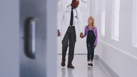 Médico-Pediatra-Masculino-Caminando-Por-El-Pasillo-Del-Hospital-Tomados-De-La-Mano-Con-Una-Paciente