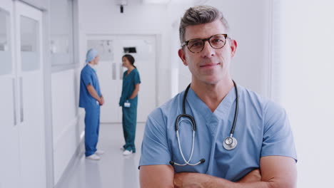 Mature-Male-Doctor-Wearing-Scrubs-Standing-In-Busy-Hospital-Corridor-With-Colleagues-In-Background