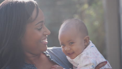 Smiling-African-American-Mother-Cuddling-And-Playing-With-Baby-Daughter-In-Garden-At-Home