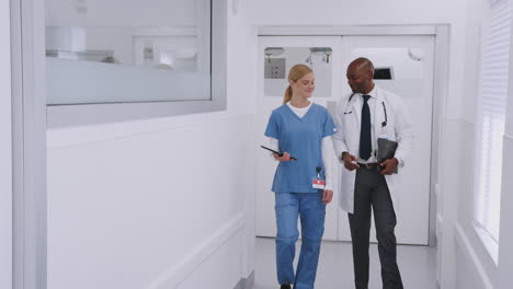 Doctor-In-White-Coat-And-Nurse-In-Scrubs-Having-Discussion-Over-Digital-Tablet-In-Hospital-Corridor