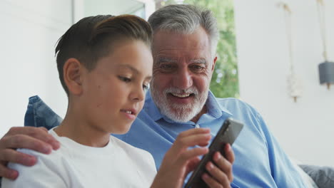 Hispanic-Grandson-Showing-Grandfather-How-To-Use-Mobile-Phone-At-Home