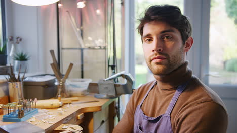 Portrait-Of-Male-Jeweller-Sitting-At-Work-Bench-Working-In-Studio