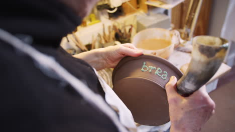 Close-Up-Of-Male-Potter-Putting-Lettering-Onto-Bread-Bowl-In-Ceramics-Studio
