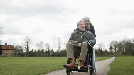 Senior-Woman-Pushing-Senior-Man-In-Wheelchair-Outdoors-In-Fall-Or-Winter-Park