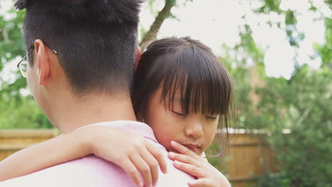 Liebevoller-Asiatischer-Vater-Kuschelt-Müde-Tochter-Im-Garten,-Während-Das-Mädchen-über-Seine-Schulter-Schaut