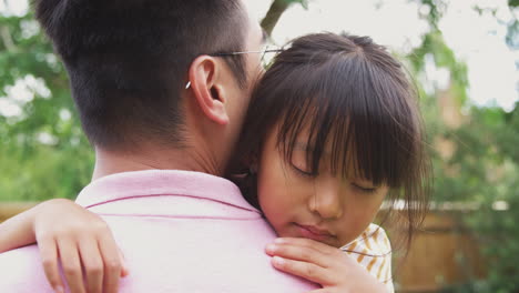 Loving-Asian-Father-Cuddling-Tired-Daughter-In-Garden-As-Girl-Looks-Over-His-Shoulder