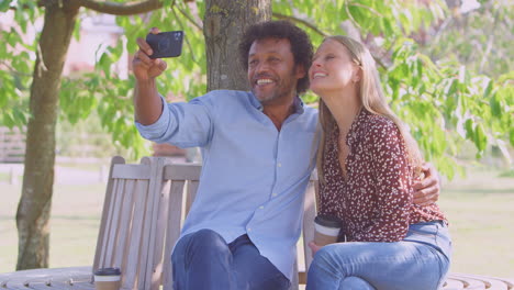 Loving-Mature-Couple-Posing-For-Selfie-On-Mobile-Phone-Sitting-On-Seat-In-Park