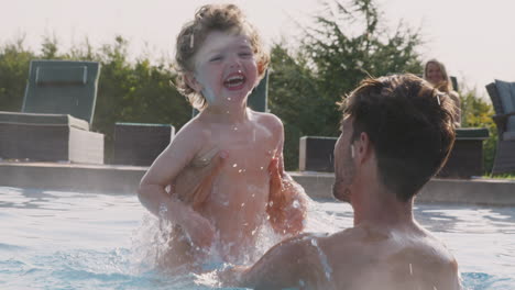Family-With-Son-And-Baby-Daughter-Having-Fun-On-Summer-Vacation-Splashing-In-Outdoor-Swimming-Pool