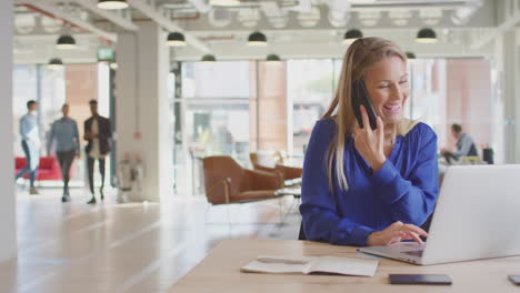Businesswoman-Sitting-At-Desk-On-Phone-Call-In-Modern-Open-Plan-Office-With-Colleagues-In-Background