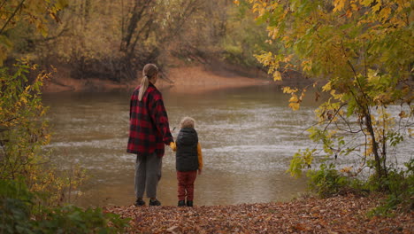 happiness-and-calmness-of-woman-with-her-little-child-at-nature-people-are-standing-on-coast-of-picturesque-forest-pond-at-autumn-day-rear-view