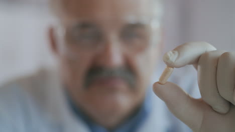 An-elderly-male-scientist-medic-holds-a-white-capsule-and-looks-at-it-extremely-close-up.-Virus-medicine.-High-quality-4k-footage