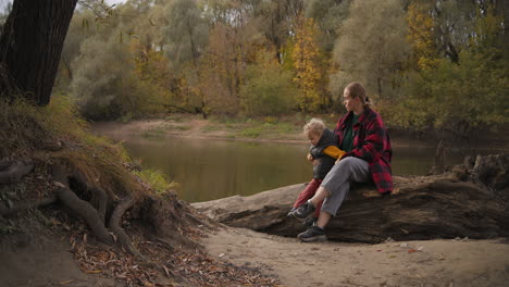 Kaukasische-Frau-Mit-Kleinem-Kind-Ruht-Sich-An-Der-Küste-Eines-Kleinen-Flusses-Im-Park-Aus-Und-Genießt-Gutes-Herbstwetter,-Glückliche-Familie-In-Der-Natur