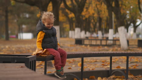 Un-Niño-Feliz-Está-Disfrutando-Del-Otoño-En-El-Parque,-Las-Hojas-Secas-Caen-Sobre-El-Bebé,-Un-Niño-Pequeño-Se-Ríe-Y-Se-Regocija-Sentado-En-Un-Banco,-Se-Divierte-Caminando-En-El-Día-De-Otoño