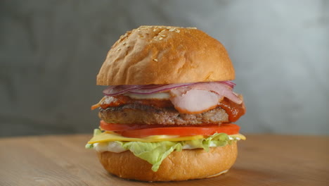 La-Hamburguesa-Gira-Sobre-Una-Tabla-De-Madera.-Una-Hamburguesa-Con-Ensalada-De-Chuleta,-Queso-Y-Tomates-Gira-Contra-Una-Pared-Gris