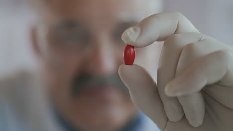 An-elderly-male-doctor-wearing-gloves-picks-up-a-red-pill-in-his-hand-looking-at-her.-Extremely-close-up.-Antibiotic-for-coronavirus-.-High-quality-4k-footage