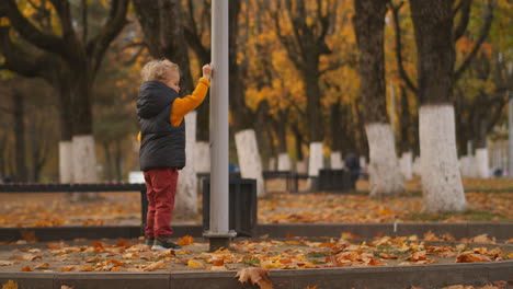 Herbstpark-In-Der-Stadt,-Kleines-Kind-Spielt-Mit-Laternenpfahl,-Steht-Allein-Auf-Der-Straße-Mit-Trockenem-Laub,-Neugieriges-Baby