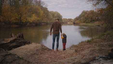 family-trip-in-woodland-father-and-little-boy-are-resting-on-coast-of-forest-lake-standing-together-and-holding-hands-rear-view-happy-childhood-with-dad
