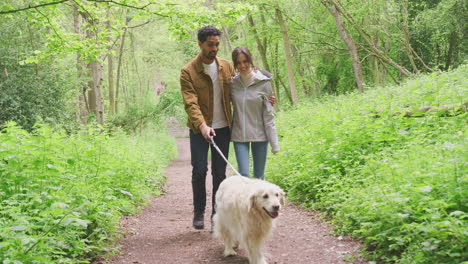 Dolly-shot-of-focus-pulled-to-young-couple-hugging-walking-along-path-through-trees-in-countryside-with-pet-golden-retriever-dog-on-leash---shot-in-slow-motion