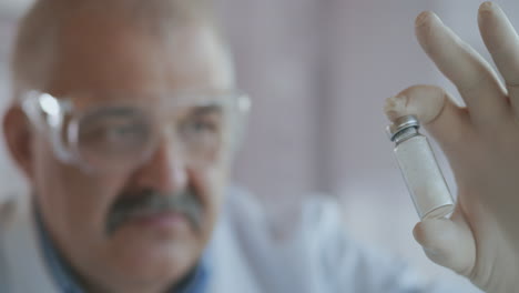 Extreme-Close-up-of-a-scientist-in-a-blue-respirator-and-protective-glasses-who-developed-a-coronavirus-vaccine-holding-an-ampoule-of-white-powder.-Narcotic-substances-and-painkillers.-High-quality-4k-footage