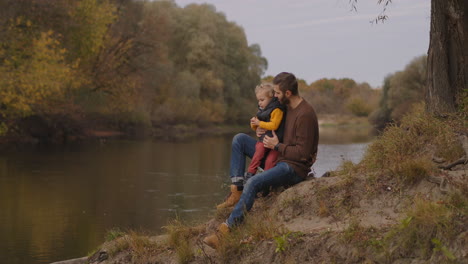 A-Principios-Del-Otoño-En-El-Bosque,-Un-Hombre-Con-Un-Hijo-Pequeño-Camina-Con-Buen-Tiempo,-Un-Hombre-Y-Un-Niño-Están-Sentados-En-La-Orilla-Del-Río,-Un-Paseo-Familiar-En-La-Naturaleza.