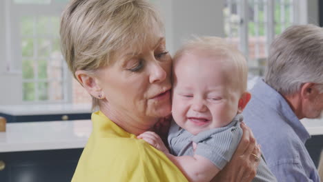 Grandmother-Cuddling-And-Comforting-Unhappy-Baby-Grandson-Sitting-At-Kitchen-Table