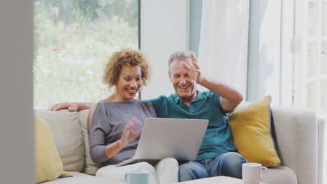 Senior-Retired-Couple-Sitting-On-Sofa-At-Home-Making-Video-Call-On-Laptop