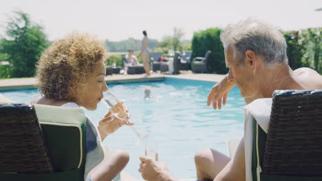 Retired-Senior-Couple-On-Loungers-Relaxing-By-Swimming-Pool-On-Summer-Vacation-Drinking-Champagne