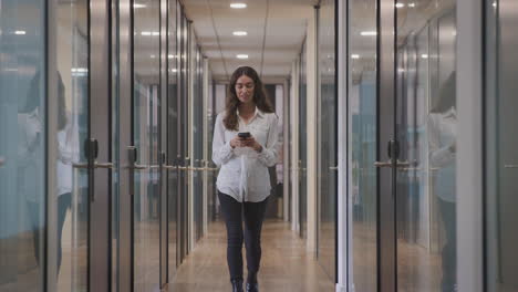 Young-Businesswoman-Walking-Along-Corridor-Of-Modern-Office-Using-Mobile-Phone