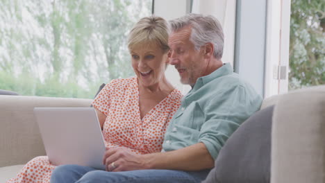 Senior-Retired-Couple-Sitting-On-Sofa-At-Home-Making-Video-Call-On-Laptop