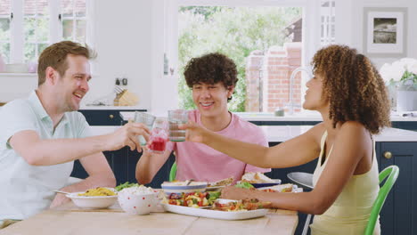 Multi-Racial-Family-With-Teenage-Son-Sitting-Around-Table-In-Kitchen-At-Home-Eating-Meal-Together