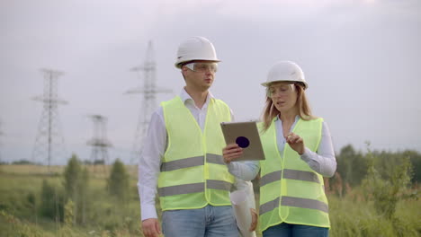 Ingeniería-Trabajando-En-Una-Torre-De-Alto-Voltaje-Verifique-La-Información-En-La-Tableta-Dos-Empleados,-Hombre-Y-Mujer