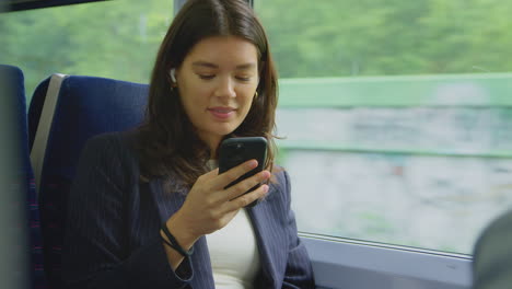 Businesswoman-With-Wireless-Earbuds-Commuting-To-Work-On-Train-Looking-At-Mobile-Phone