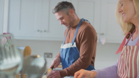 Mature-couple-wearing-aprons-preparing-ingredients-and-cooking-meal-at-home-together---shot-in-slow-motion