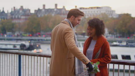 Couple-in-autumn-or-fall-meeting-on-date-in-London-by-river-Thames-with-man-giving-woman-gift-and-bouquet-of-flowers---shot-in-slow-motion