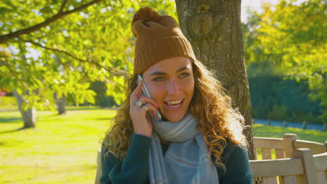Smiling-Young-Woman-Wearing-Hat-And-Scarf-Sitting-On-Bench-In-Autumn-Park-Talking-On-Mobile-Phone