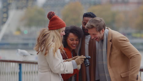 Group-of-friends-outdoors-wearing-coats-and-scarves-looking-at-photos-on-camera-on-autumn-or-winter-trip-to-London---shot-in-slow-motion