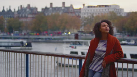 Woman-wearing-winter-coat-leaning-on-railings-by-river-Thames-in-London-at-dusk---shot-in-slow-motion