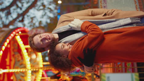 Vertical-video-of-couple-enjoying-on-date-in-standing-by-carousel-at-Christmas-funfair-at-night---shot-in-slow-motion