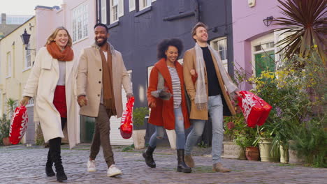 Two-multi-cultural-couples-arm-in-arm-as-they-walk-along-cobbled-mews-street-on-visit-to-city-in-autumn-or-winter-carrying-sale-shopping-bags---shot-in-slow-motion